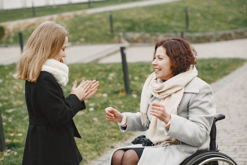 A woman who uses a wheelchair laughing and playing with a young girl outdoors